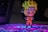 A small figurine of an anime character stands confidently with arms crossed, wearing an orange jumpsuit and a headband with a unique symbol. The background features a colorful, illuminated surface with a blend of purple and blue hues.