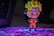 A small figurine of an anime character stands confidently with arms crossed, wearing an orange jumpsuit and a headband with a unique symbol. The background features a colorful, illuminated surface with a blend of purple and blue hues.