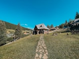 A wooden house with a sloped roof sits on a gently sloping grassy hill, surrounded by trees and a wooden fence. A stone path leads to the house, and there are benches and a wagon wheel nearby. In the background, a lush forest stretches into the distance under a clear blue sky.