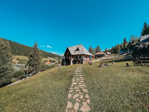 A wooden house with a sloped roof sits on a gently sloping grassy hill, surrounded by trees and a wooden fence. A stone path leads to the house, and there are benches and a wagon wheel nearby. In the background, a lush forest stretches into the distance under a clear blue sky.