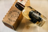 A clear glass bottle with a dark label featuring vintage-style text lies next to a brown cardboard box. The bottle contains a light amber liquid and has a dark brown cap. The cardboard box has printed text resembling a stamp, which includes numbers and letters indicating a product made in 1933.