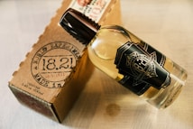 A clear glass bottle with a dark label featuring vintage-style text lies next to a brown cardboard box. The bottle contains a light amber liquid and has a dark brown cap. The cardboard box has printed text resembling a stamp, which includes numbers and letters indicating a product made in 1933.
