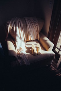 A cozy armchair is bathed in soft, natural light from a nearby window. Draped across the chair is a light-colored, flowing fabric, perhaps a scarf or shawl. A pair of beige, open-toed slippers are neatly placed on the seat, adding to the sense of tranquility and comfort.