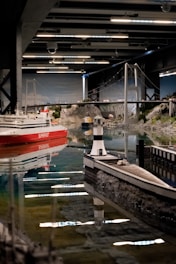 A detailed model display featuring a miniature bridge, a red and white ship, rocky terrain, and a lighthouse. The scene is set indoors with a ceiling structure visible above. The water reflects the models, enhancing the lifelike appearance.