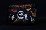 A black case featuring various space-themed designs, including a NASA logo, an astronaut, a rocket, planets, and cryptic text in bright colors such as orange and white. The artwork combines elements of space exploration with graffiti-like writing.