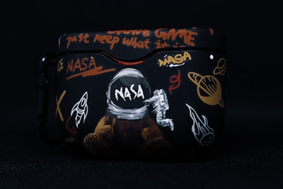 A black case featuring various space-themed designs, including a NASA logo, an astronaut, a rocket, planets, and cryptic text in bright colors such as orange and white. The artwork combines elements of space exploration with graffiti-like writing.