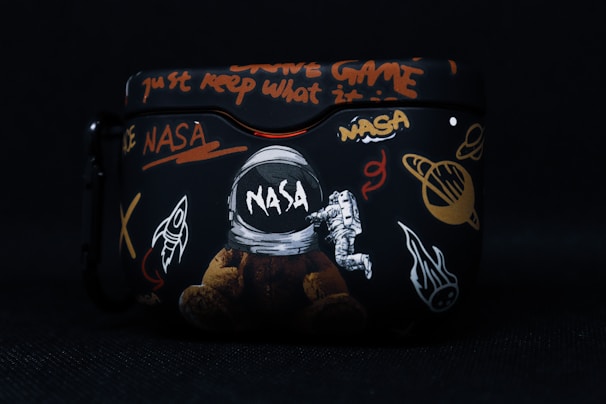 A black case featuring various space-themed designs, including a NASA logo, an astronaut, a rocket, planets, and cryptic text in bright colors such as orange and white. The artwork combines elements of space exploration with graffiti-like writing.