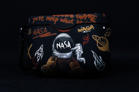 A black case featuring various space-themed designs, including a NASA logo, an astronaut, a rocket, planets, and cryptic text in bright colors such as orange and white. The artwork combines elements of space exploration with graffiti-like writing.