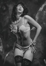A person is posing in lingerie with a retro-style aesthetic. The image is in black and white, emphasizing the contrasting textures of lace and pearls. The background appears to be a textured wall that adds depth to the composition.