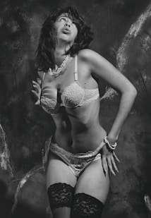 A person is posing in lingerie with a retro-style aesthetic. The image is in black and white, emphasizing the contrasting textures of lace and pearls. The background appears to be a textured wall that adds depth to the composition.