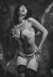 A person is posing in lingerie with a retro-style aesthetic. The image is in black and white, emphasizing the contrasting textures of lace and pearls. The background appears to be a textured wall that adds depth to the composition.