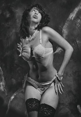 A person is posing in lingerie with a retro-style aesthetic. The image is in black and white, emphasizing the contrasting textures of lace and pearls. The background appears to be a textured wall that adds depth to the composition.
