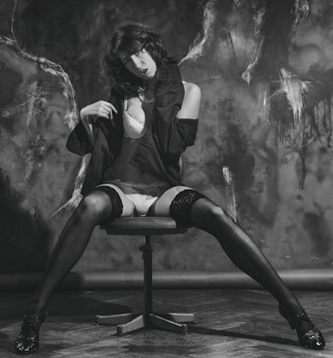 A person is sitting on a swivel chair with legs spread on a wooden floor. Wearing a black jacket over lingerie, including stockings and high heels. The background features textured dark patterns, creating a dramatic contrast.