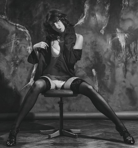 A person is sitting on a swivel chair with legs spread on a wooden floor. Wearing a black jacket over lingerie, including stockings and high heels. The background features textured dark patterns, creating a dramatic contrast.