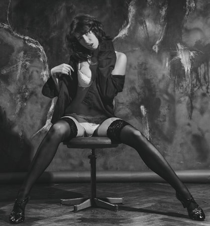 A person is sitting on a swivel chair with legs spread on a wooden floor. Wearing a black jacket over lingerie, including stockings and high heels. The background features textured dark patterns, creating a dramatic contrast.