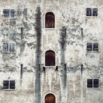 A weathered stucco wall features three vertically aligned wooden doors with arched tops, flanked by several small, shuttered windows. The wall displays a rough texture and dark streaks, indicating age and exposure to the elements.