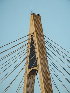 A modern cable-stayed bridge tower with numerous cables extending outward. The structure is made of concrete and steel, showcasing an architectural design with vertical and diagonal elements.
