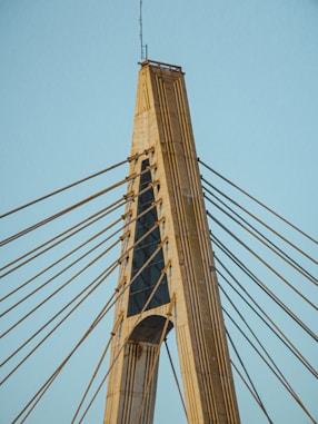 A modern cable-stayed bridge tower with numerous cables extending outward. The structure is made of concrete and steel, showcasing an architectural design with vertical and diagonal elements.
