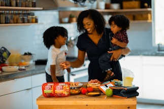A woman is in a kitchen holding a young child, with another child standing next to her. They appear to be preparing a meal or snack together, surrounded by food items and containers on the counter. The setting is bright and inviting with shelves of spices and kitchen tools in the background.