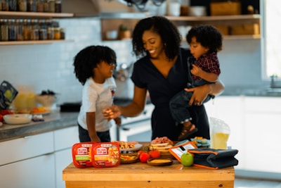 A woman is in a kitchen holding a young child, with another child standing next to her. They appear to be preparing a meal or snack together, surrounded by food items and containers on the counter. The setting is bright and inviting with shelves of spices and kitchen tools in the background.