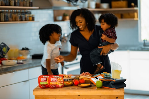 A woman is in a kitchen holding a young child, with another child standing next to her. They appear to be preparing a meal or snack together, surrounded by food items and containers on the counter. The setting is bright and inviting with shelves of spices and kitchen tools in the background.