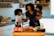 A woman is in a kitchen holding a young child, with another child standing next to her. They appear to be preparing a meal or snack together, surrounded by food items and containers on the counter. The setting is bright and inviting with shelves of spices and kitchen tools in the background.