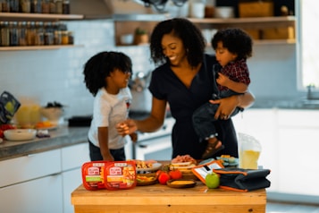A woman is in a kitchen holding a young child, with another child standing next to her. They appear to be preparing a meal or snack together, surrounded by food items and containers on the counter. The setting is bright and inviting with shelves of spices and kitchen tools in the background.