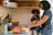A woman is preparing food in a kitchen while smiling at a child sitting on the counter. The child looks playful and is holding a small piece of food. The kitchen has light-colored tiles and various kitchen utensils are organized on the counter. A basket with bananas and other food items is visible in the background.