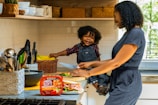 A mother and child are in a kitchen, with the child sitting on the counter. The mother is cutting vegetables on a wooden board, while the child happily holds a knife. The kitchen has white tiled walls, with a collection of knives, bowls, and a basket of bananas visible. The atmosphere is bright and cheerful, with natural light coming through a window.