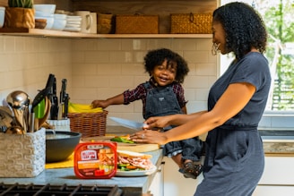 A mother and child are in a kitchen, with the child sitting on the counter. The mother is cutting vegetables on a wooden board, while the child happily holds a knife. The kitchen has white tiled walls, with a collection of knives, bowls, and a basket of bananas visible. The atmosphere is bright and cheerful, with natural light coming through a window.