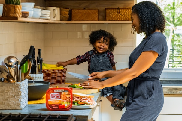 A mother and child are in a kitchen, with the child sitting on the counter. The mother is cutting vegetables on a wooden board, while the child happily holds a knife. The kitchen has white tiled walls, with a collection of knives, bowls, and a basket of bananas visible. The atmosphere is bright and cheerful, with natural light coming through a window.
