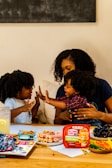 A family scene with a woman and two children enjoying a playful moment at a table. The table is filled with snacks including rolled-up deli meats, a container labeled 'Hillshire Farm', grapes, and a variety of colorful vegetables. The atmosphere is warm and familial, with the woman and children displaying affectionate gestures.