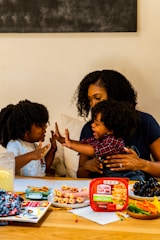 A family scene with a woman and two children enjoying a playful moment at a table. The table is filled with snacks including rolled-up deli meats, a container labeled 'Hillshire Farm', grapes, and a variety of colorful vegetables. The atmosphere is warm and familial, with the woman and children displaying affectionate gestures.