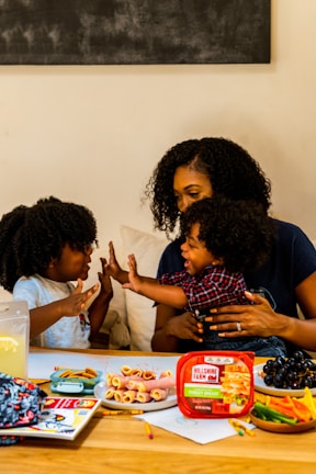 A family scene with a woman and two children enjoying a playful moment at a table. The table is filled with snacks including rolled-up deli meats, a container labeled 'Hillshire Farm', grapes, and a variety of colorful vegetables. The atmosphere is warm and familial, with the woman and children displaying affectionate gestures.