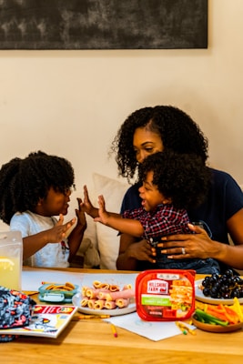 A family scene with a woman and two children enjoying a playful moment at a table. The table is filled with snacks including rolled-up deli meats, a container labeled 'Hillshire Farm', grapes, and a variety of colorful vegetables. The atmosphere is warm and familial, with the woman and children displaying affectionate gestures.