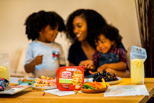 A happy family scene at a dining table, featuring a woman and two children sharing a moment together. The table is filled with a variety of food items, including a package of Hillshire Farm turkey breast, a plate of black grapes, and colorful sliced vegetables. Two pouches of lemonade with lemon slices sit on either side of the table. Children's drawings and crayons are visible on the table.