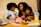 A happy family scene at a dining table, featuring a woman and two children sharing a moment together. The table is filled with a variety of food items, including a package of Hillshire Farm turkey breast, a plate of black grapes, and colorful sliced vegetables. Two pouches of lemonade with lemon slices sit on either side of the table. Children's drawings and crayons are visible on the table.