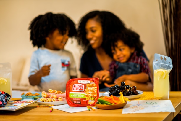 A happy family scene at a dining table, featuring a woman and two children sharing a moment together. The table is filled with a variety of food items, including a package of Hillshire Farm turkey breast, a plate of black grapes, and colorful sliced vegetables. Two pouches of lemonade with lemon slices sit on either side of the table. Children's drawings and crayons are visible on the table.
