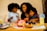 A happy family scene at a dining table, featuring a woman and two children sharing a moment together. The table is filled with a variety of food items, including a package of Hillshire Farm turkey breast, a plate of black grapes, and colorful sliced vegetables. Two pouches of lemonade with lemon slices sit on either side of the table. Children's drawings and crayons are visible on the table.