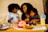 A happy family scene at a dining table, featuring a woman and two children sharing a moment together. The table is filled with a variety of food items, including a package of Hillshire Farm turkey breast, a plate of black grapes, and colorful sliced vegetables. Two pouches of lemonade with lemon slices sit on either side of the table. Children's drawings and crayons are visible on the table.
