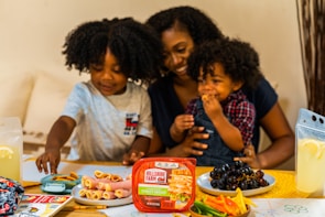 A family scene with two young children and an adult, engaging in a meal preparation or snack time. Various food items, including a package of turkey breast, rolled cold cuts, grapes, sliced bell peppers, and lemonade, are arranged on a table. The atmosphere is warm and nurturing, with the adult appearing to assist or supervise. The children are smiling and focused on the food. Art supplies and drawings are visible, suggesting a playful environment.