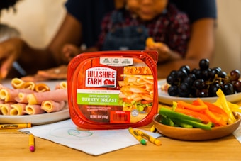 A kitchen table displays an assortment of snacks, including a package of Hillshire Farm turkey breast prominently positioned at the front. Surrounding it are plates with rolled slices of ham, a platter of colorful sliced bell peppers, baby carrots, and a bowl of dark grapes. Crayons and a child's drawing are on the table, suggesting a family activity. Two people, possibly a parent and child, are visible in the background engaging in meal preparation.