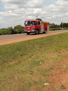 A red fire truck is driving along a rural road bordered by green grass and trees. The sky is filled with scattered clouds, and there are telegraph poles lining the road.