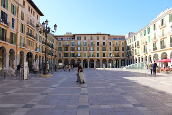 A spacious plaza surrounded by tall, beige-colored buildings with arched windows and green shutters. The foreground features a patterned cobblestone pavement, and several people are walking or sitting at outdoor tables under umbrellas. There is a classic street lamp in the center of the square beneath a clear blue sky.