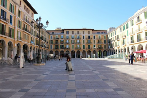 A spacious plaza surrounded by tall, beige-colored buildings with arched windows and green shutters. The foreground features a patterned cobblestone pavement, and several people are walking or sitting at outdoor tables under umbrellas. There is a classic street lamp in the center of the square beneath a clear blue sky.
