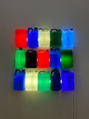 A grid of illuminated plastic jugs hangs on a wall, each displaying vibrant colors such as red, blue, green, and white. The jugs are arranged in three horizontal rows and five vertical columns, with internal lighting giving them a glowing effect.
