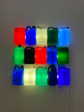 A grid of illuminated plastic jugs hangs on a wall, each displaying vibrant colors such as red, blue, green, and white. The jugs are arranged in three horizontal rows and five vertical columns, with internal lighting giving them a glowing effect.