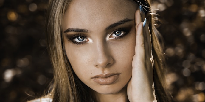A close-up portrait of a person with striking blue eyes and long eyelashes. The person has long, light brown hair and is touching their face with carefully manicured nails. The background is softly blurred with hints of brown and gold, creating a bokeh effect.