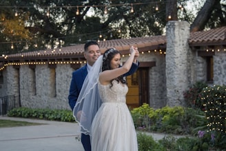 A couple is smiling and dancing together in an outdoor setting, surrounded by greenery and festive string lights. The bride wears a white dress and veil, and the groom is in a blue suit. The background features a stone building with a tiled roof.