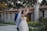 A couple is smiling and dancing together in an outdoor setting, surrounded by greenery and festive string lights. The bride wears a white dress and veil, and the groom is in a blue suit. The background features a stone building with a tiled roof.